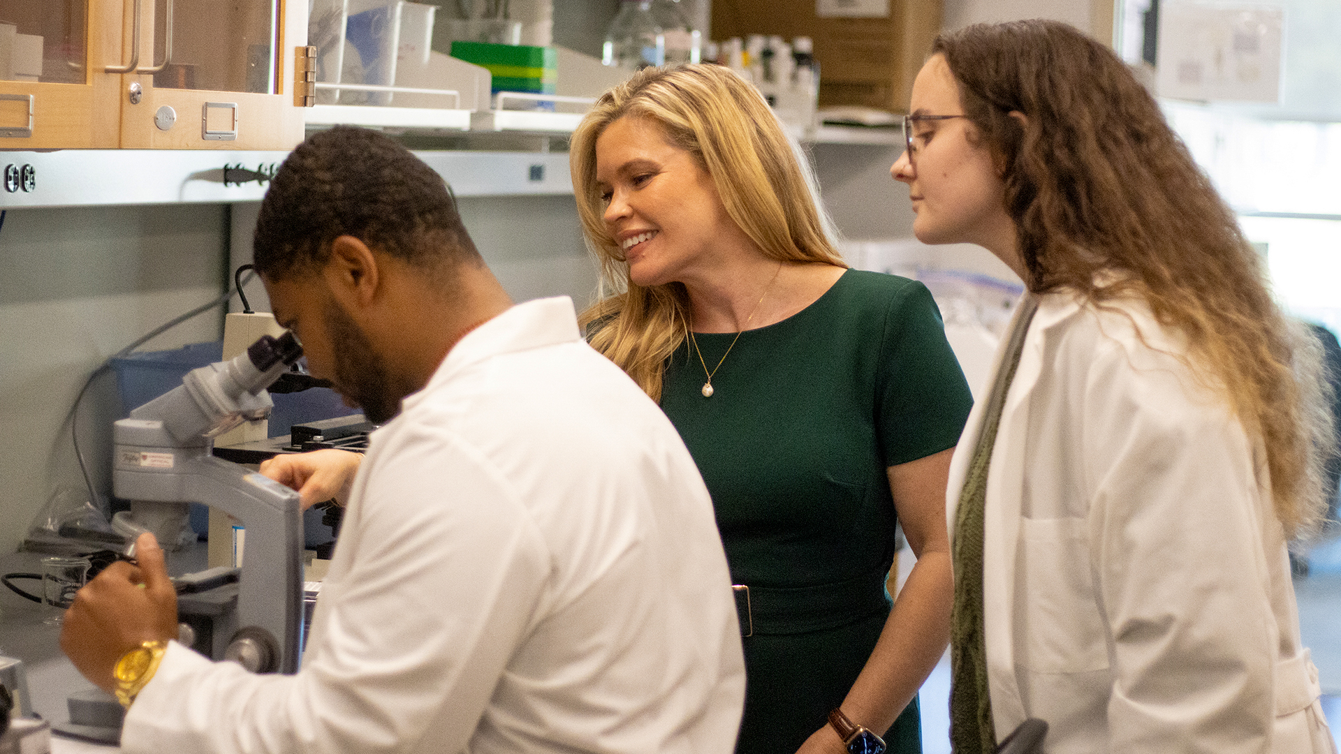 Professor stands with students at microscope